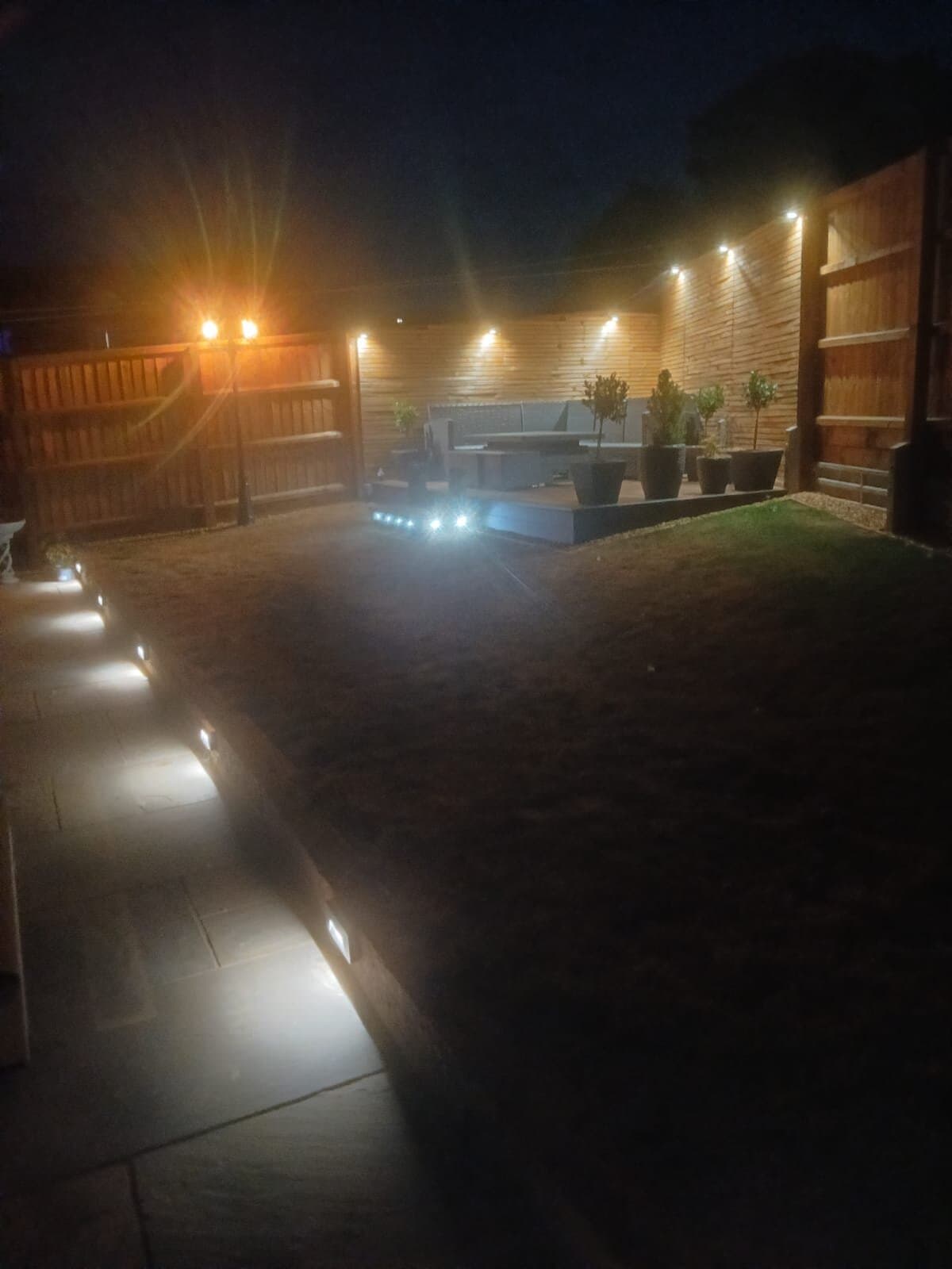 Garden at night lit by LED deck lights set in the path and lights along the fence